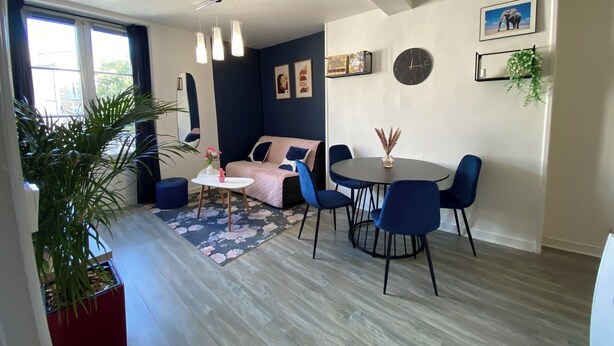 A modern living room with a sofa, a round table, and chairs. There is a large window, a plant, and a clock on the wall. - L'Indigo, charming T2 10 min from the center, Netflix Prime (Poitiers)