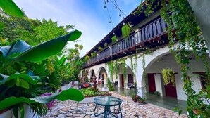 An outdoor courtyard with a stone floor, a glass table, and chairs. There are arches and a balcony with hanging plants. - GARDEN HOTEL (Copan Ruinas)
