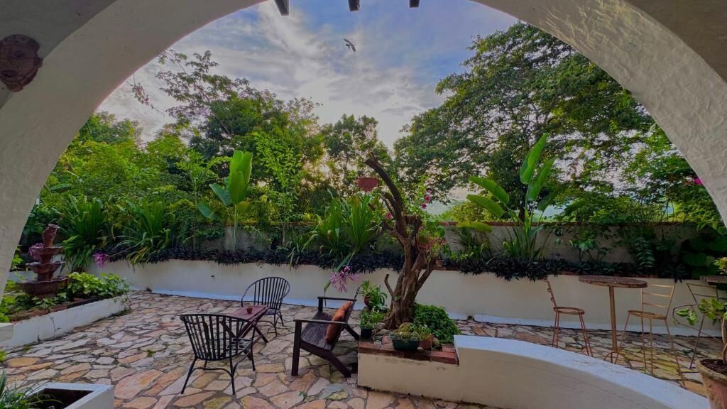 A covered patio with stone flooring, a small table, and chairs, surrounded by lush greenery and a tree.