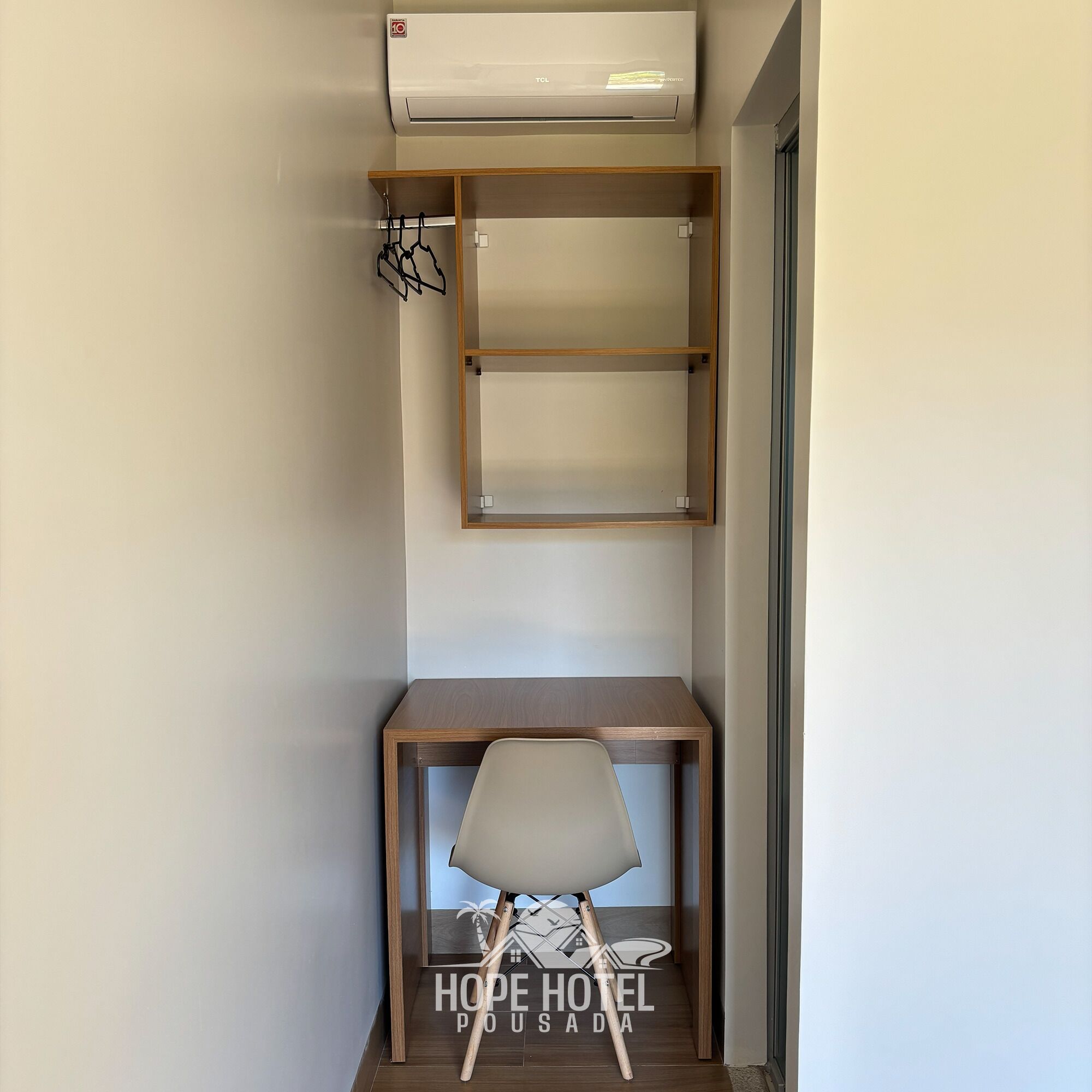 A compact hotel room with a built-in desk and chair, a wall-mounted shelf, and an air conditioning unit.