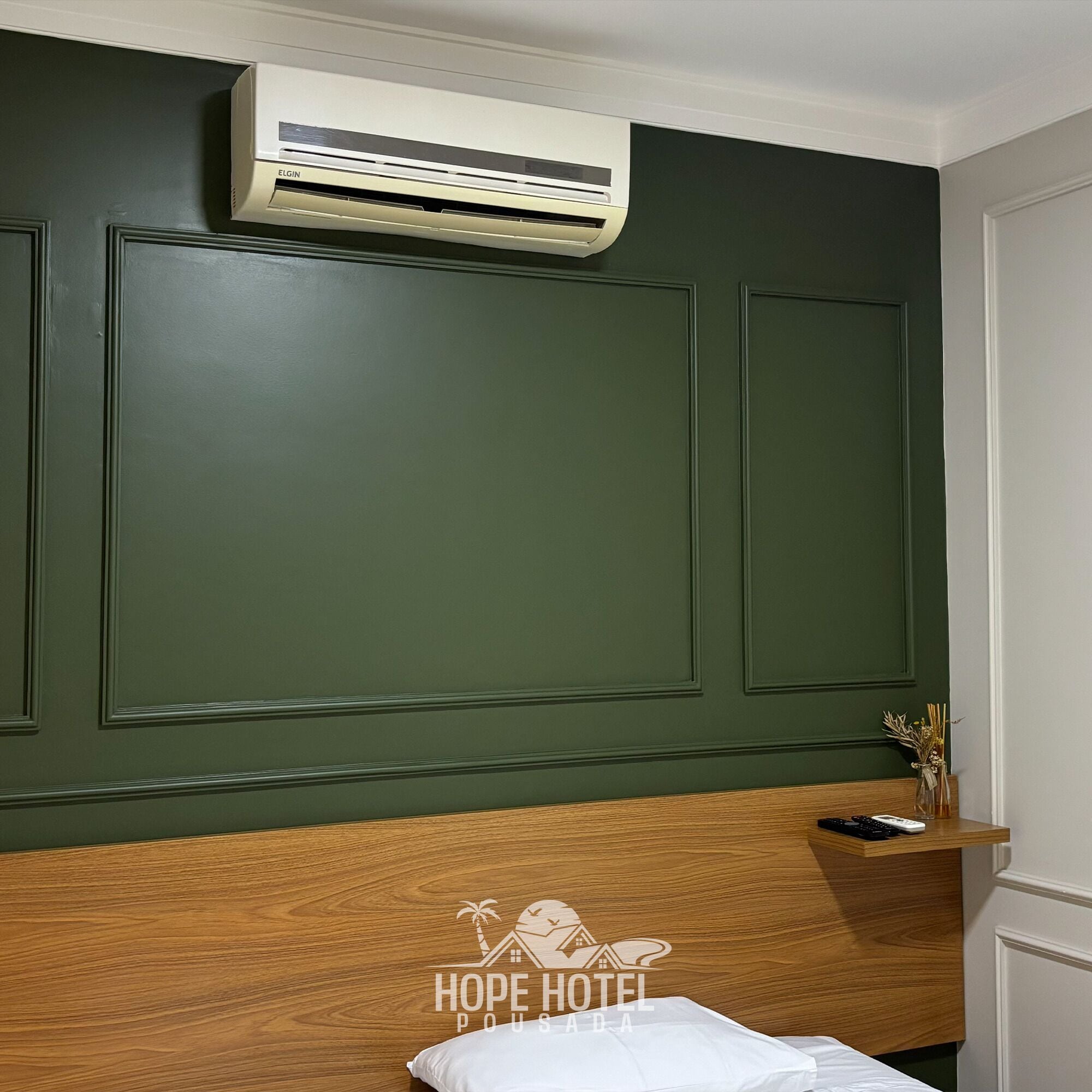 Habitación de hotel con una cama, cabecera de madera, aire acondicionado de pared y una pequeña estantería con un control remoto y un jarrón de flores secas.
