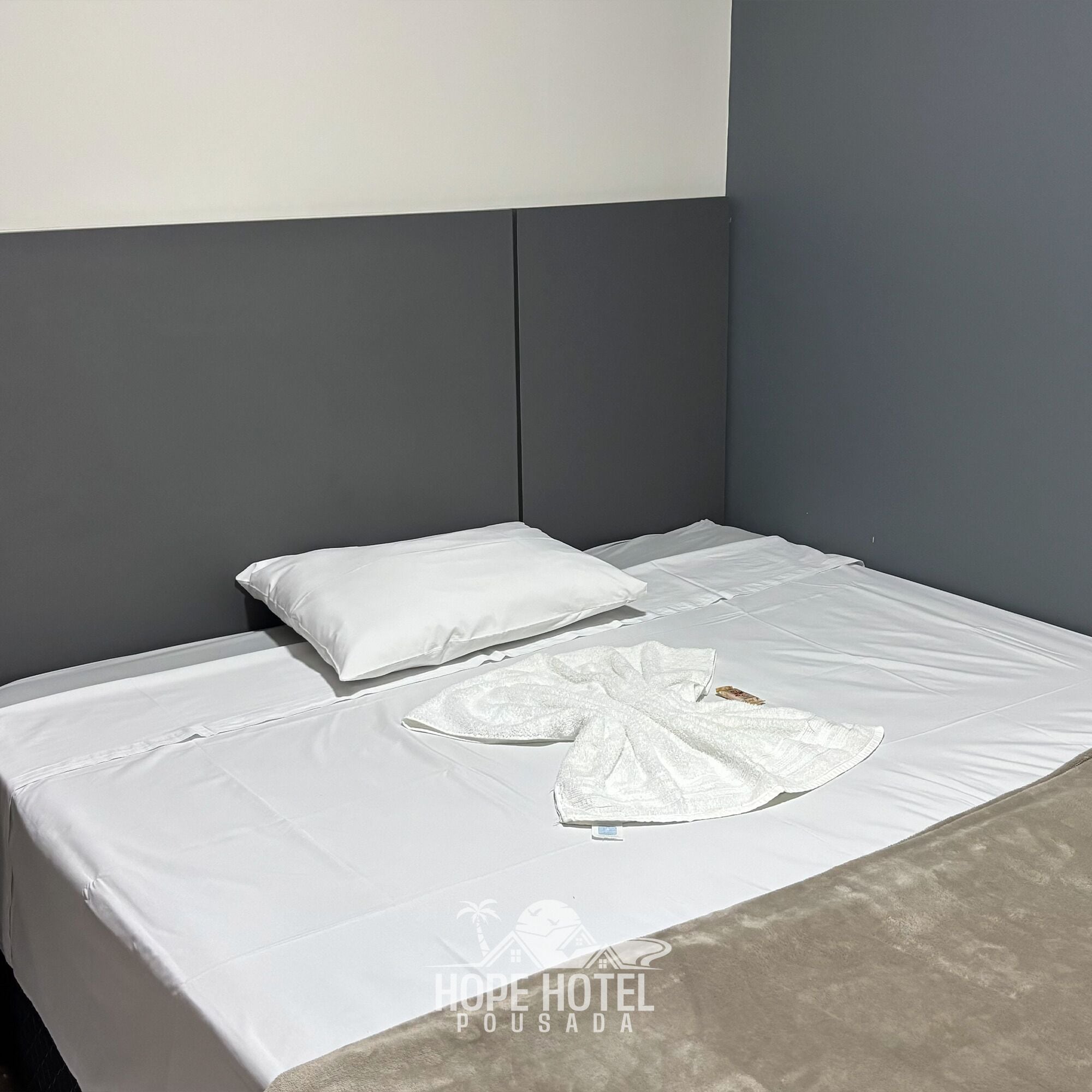 A neatly made bed with white linens and pillows, a grey headboard, and a logo for Hope Hotel Pousada.
