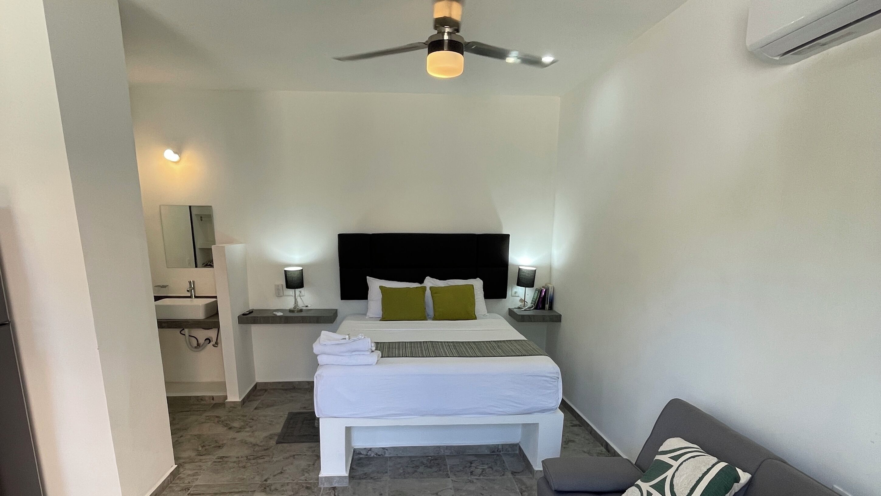 Habitación de hotel con cama, mesitas de noche, espejo, ventilador de techo y baño visible a través de una puerta abierta.