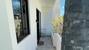 Un estrecho pasillo exterior con una pared blanca, una puerta negra y una pequeña silla blanca.