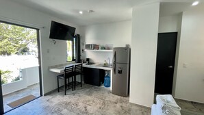 Una cocina compacta con una televisión de pantalla plana, un microondas y un refrigerador. Hay una peque ña zona de bar con dos sillas. La cocina tiene un ventanal con vistas a árboles y una puerta que conduce a otra habitación.