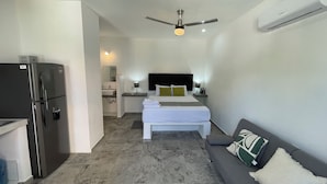Una habitación de hotel compacta con cama, un pequeño rincón de descanso, kitchenette y ventilador de techo.