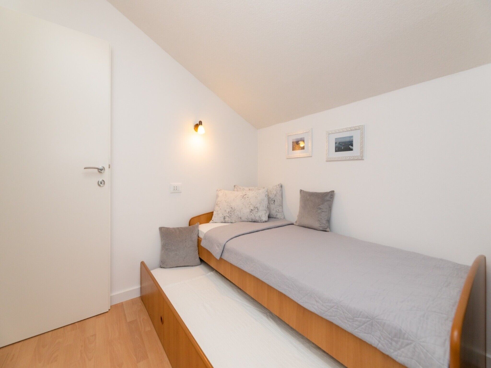 A small, single-bed room with a wooden headboard, a bedside table, and a wall-mounted light fixture. There are two framed pictures on the wall.