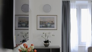 Ett modernt vardagsrum med en platt-TV, ett runt soffbord i trä, en vas med blommor och ett stort fönster med gardiner.