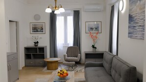 Ett modernt vardagsrum med en grå soffa, ett träbord med en fruktskål, en grå fåtölj och ett runt träbord. Det finns ett grått skåp med lådor, en grå hylla och en grå matta på golvet.