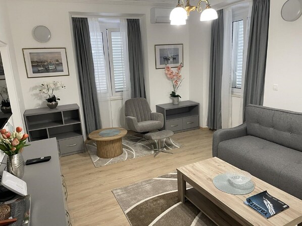 En moderne stue med en grå sofa, et rundt sofabord og en grå lænestol. To vinduer med grå gardiner, et lille sofabord med en blomstervase, og en reol.