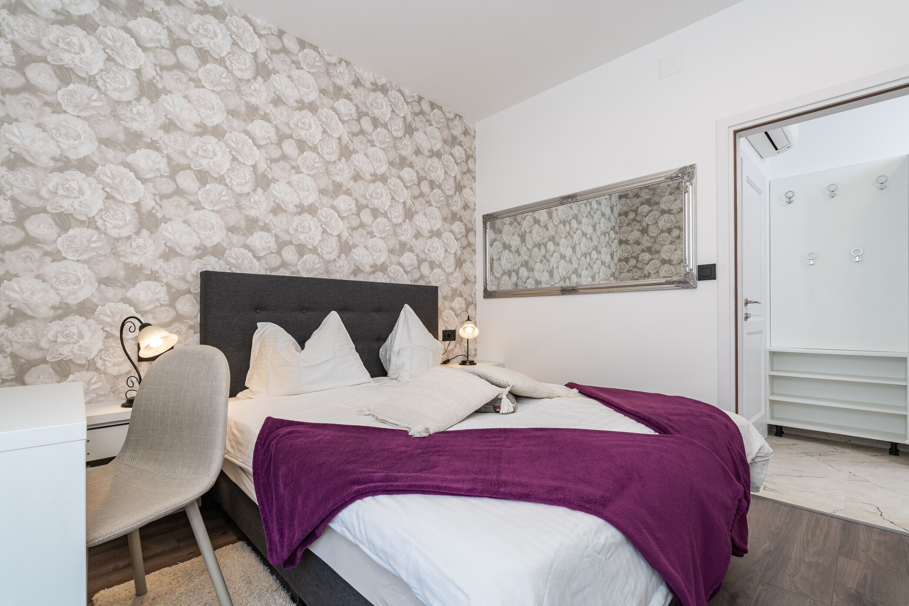 Ein Schlafzimmer mit einem Bett, einem Sessel, einem Schreibtisch, einem Kleiderschrank und einem Spiegel.