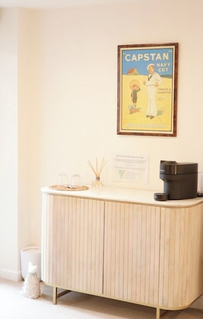 Ein Zimmer mit einem Holzkasten, einem gerahmten Poster, einer Kaffeemaschine und einem kleinen Tisch mit Glasbehältern.
