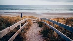 Ein hölzerner Weg führt zu einem Strand, im Hintergrund das Meer.