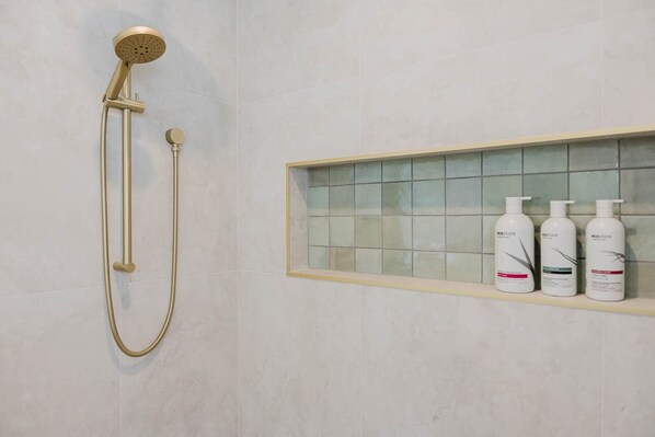 Eine Dusche mit einem goldfarbenen Brausekopf und einer Handbrause, eine geflieste Wand und ein Ablagefach mit drei Lotionflaschen.