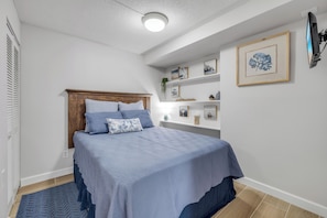A bedroom with a bed, a headboard, a bedside table, a wall-mounted TV, and a shelf with decorative items. - Cozy Coastal Retreat–1BR w/Kitchen & Ocean Access-unit 9 (New Smyrna Beach)