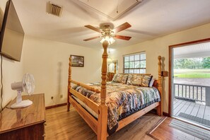 Un dormitorio con cama de madera, ventilador de techo, un ventilador sobre una mesa de madera y vistas al exterior a través de un ventanal.
