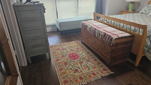 Un dormitorio con cómoda de madera, una banca, cama con colcha estampada, mesita de noche con lámpara y ventana con persianas.