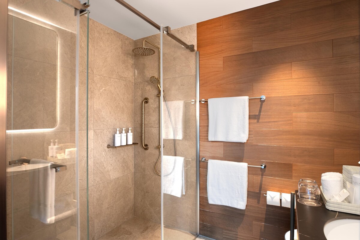 A modern bathroom with a glass-enclosed shower, wooden wall paneling, a glass partition, and a vanity with a mirror and towel rack.