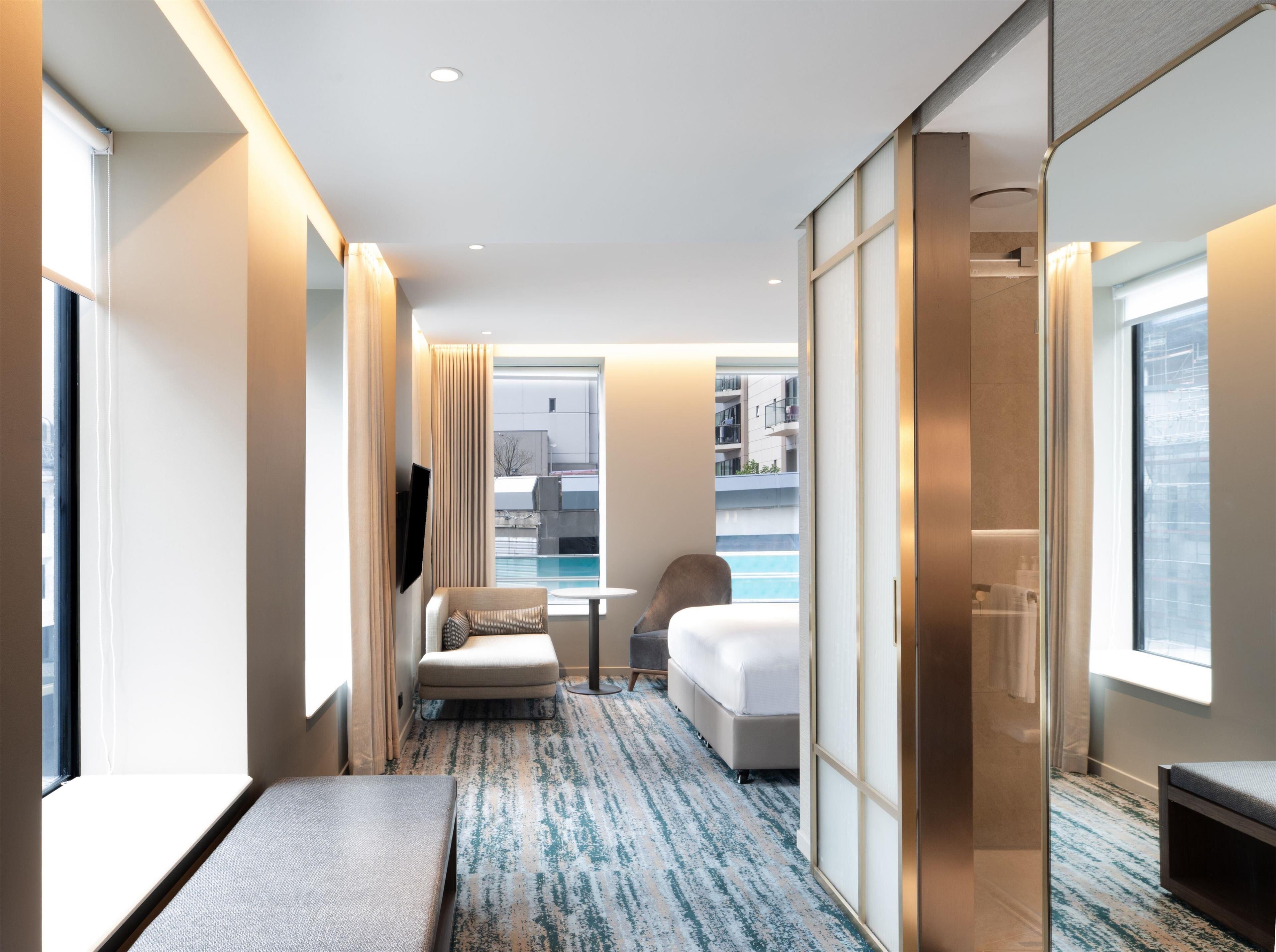 A modern hotel room with a bed, a seating area with a chair and a small table, a large window with a view of the city, and a pool visible through the window.