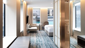 A modern hotel room with a bed, a seating area with a chair and a small table, a large window with a view of the city, and a pool visible through the window. - Doubletree By Hilton Auckland Albert Street (Auckland)