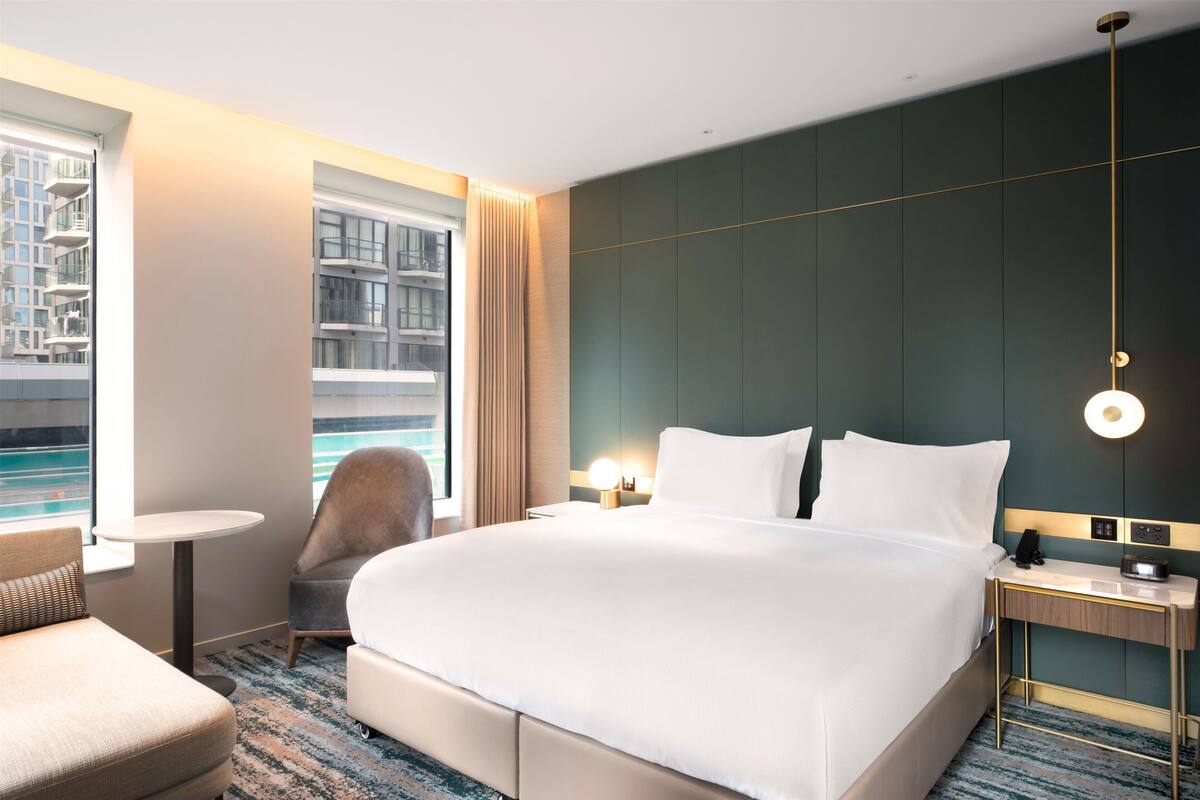 A hotel room with a large bed, a small table, a chair, a bedside table with a lamp, and a view of a building through the window.