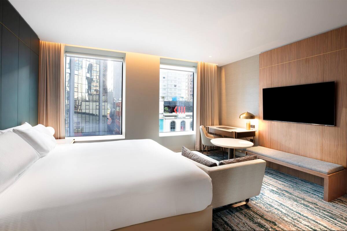 A hotel room with a large bed, a desk, a chair, a TV, and a view of the city.