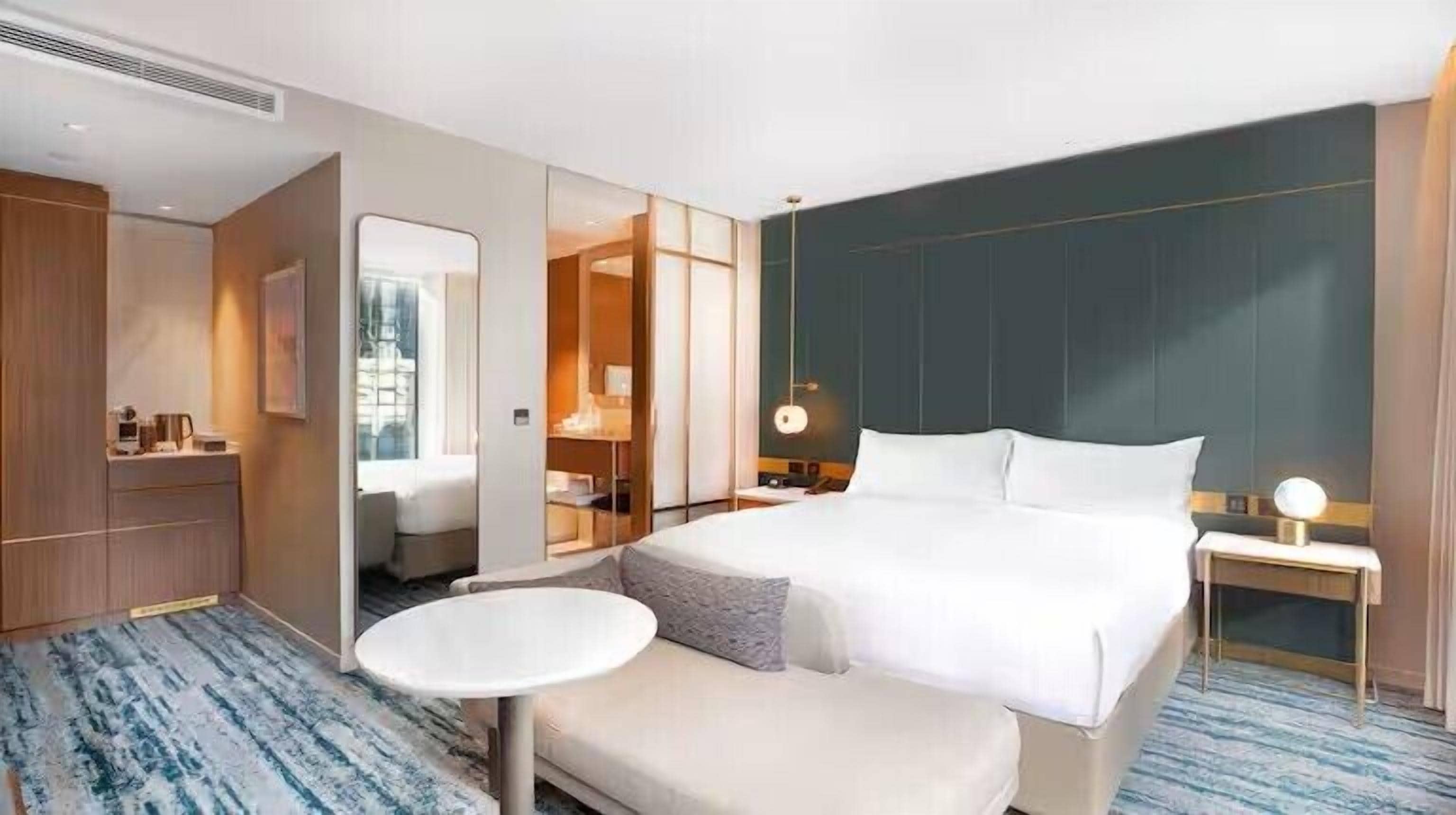 A hotel room with a large bed, a small round table, a bedside lamp, and a view of the city through the window.