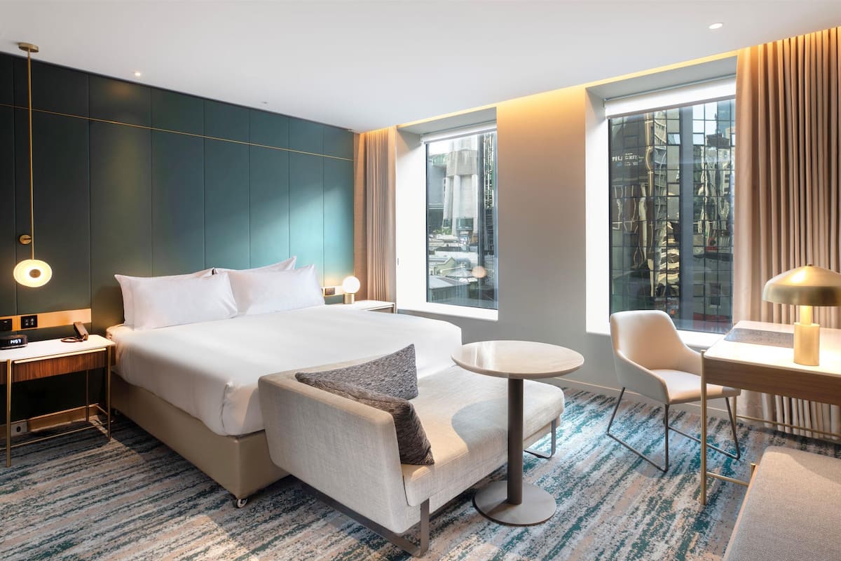 A modern hotel room with a large bed, a round table, a chair, and a desk with a lamp.