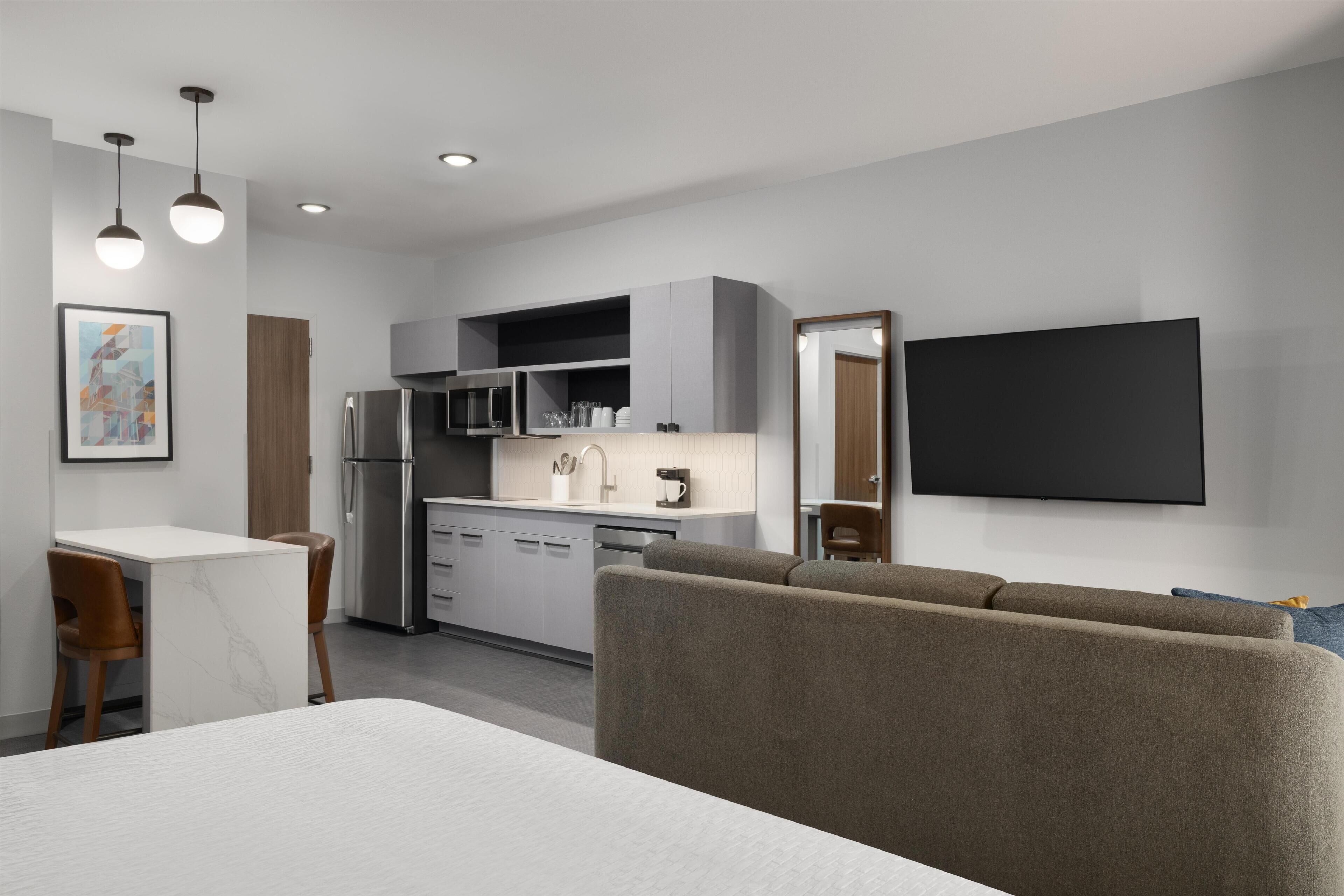 A modern kitchen with a refrigerator, microwave, and coffee maker, a dining area with a table and chairs, a sofa, and a TV mounted on the wall.