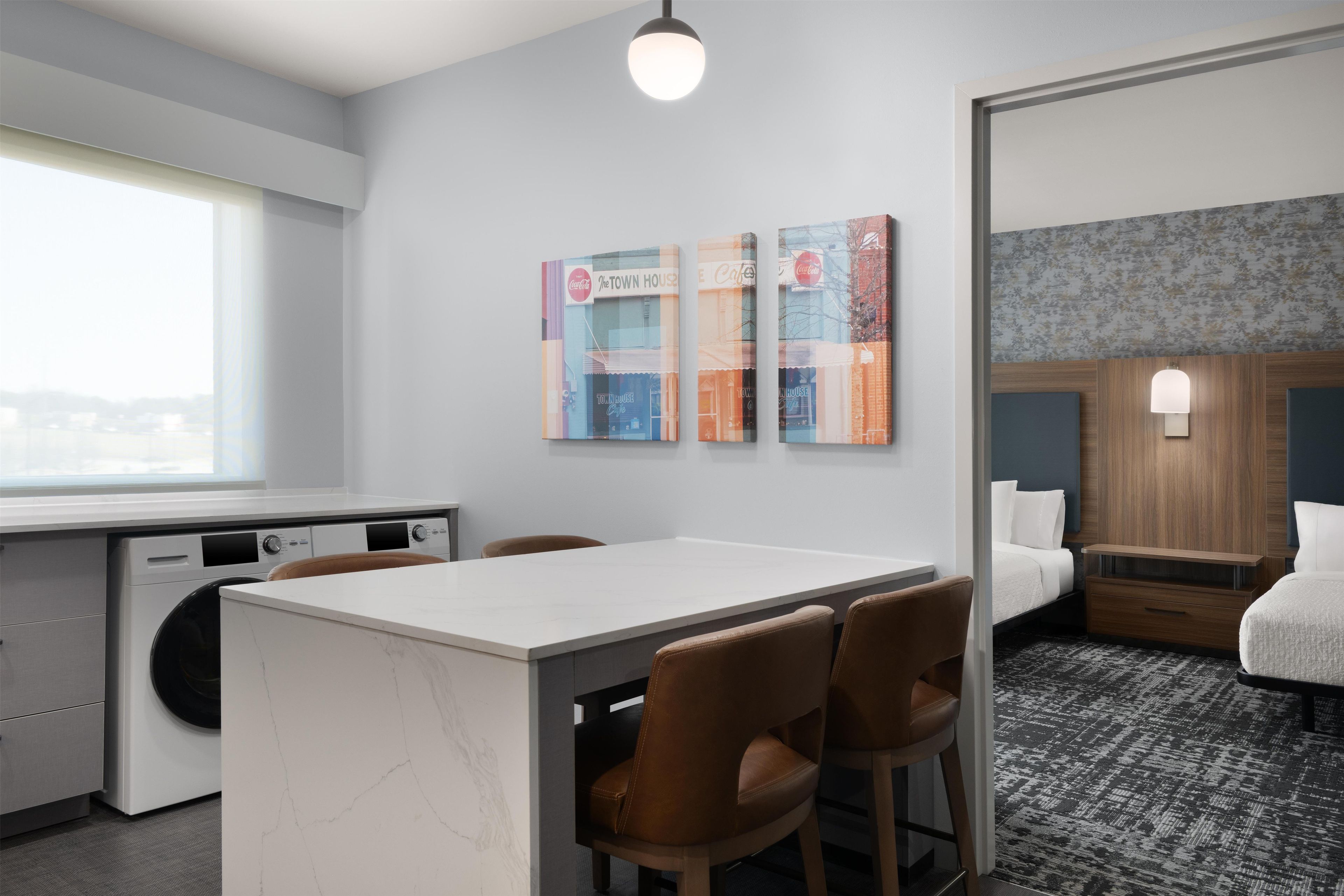 A hotel room with a kitchenette, a dining area with a table and chairs, a bed with a wooden headboard, and a wall-mounted light fixture.