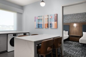 Habitación de hotel con kitchenette, zona de comedor con mesa y sillas, cama con cabecera de madera y lámpara fijada a la pared.