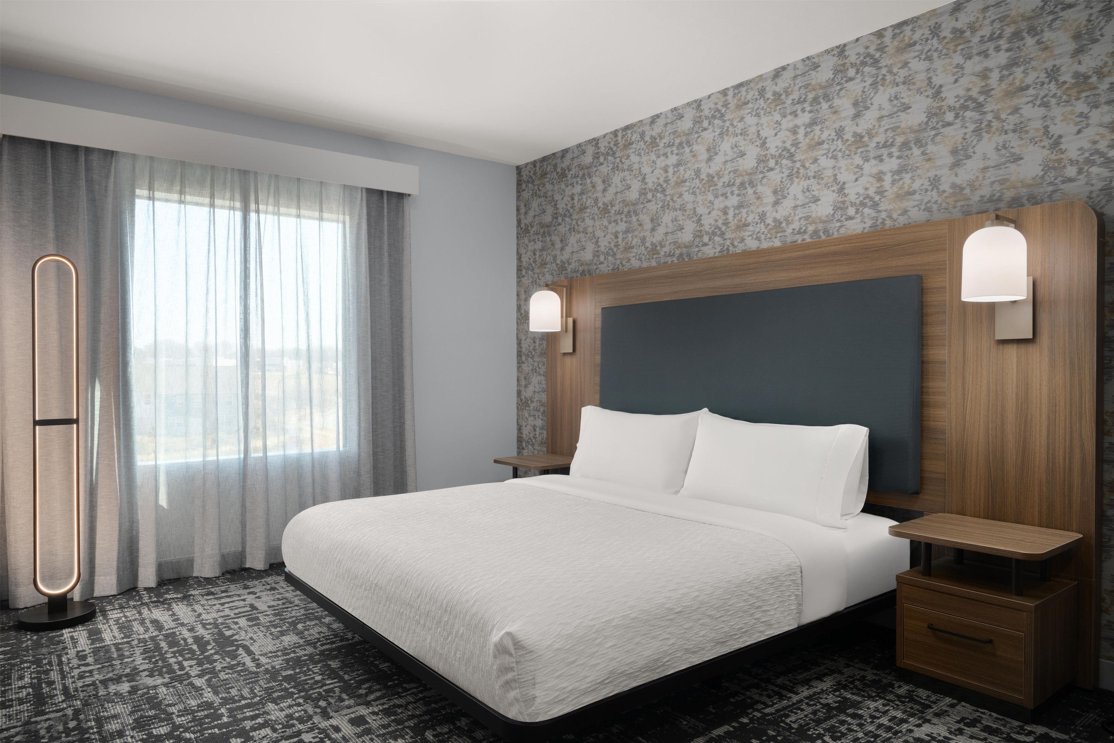 Habitación de hotel con una cama grande, una mesita de noche, una lámpara y un ventanal con cortinas.