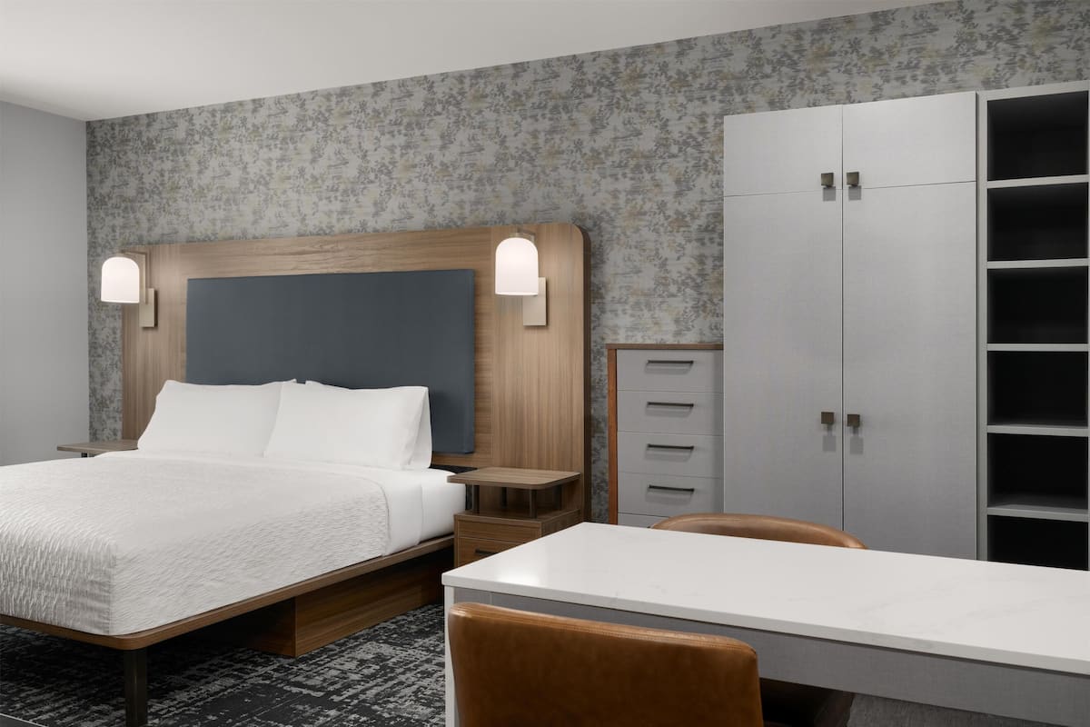 A hotel room with a bed, bedside tables, a desk, a chair, a wardrobe, and a wall with a floral pattern.