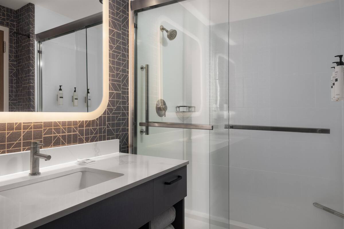 A modern bathroom with a glass shower enclosure, a vanity with a sink, and a mirror with a geometric backsplash.
