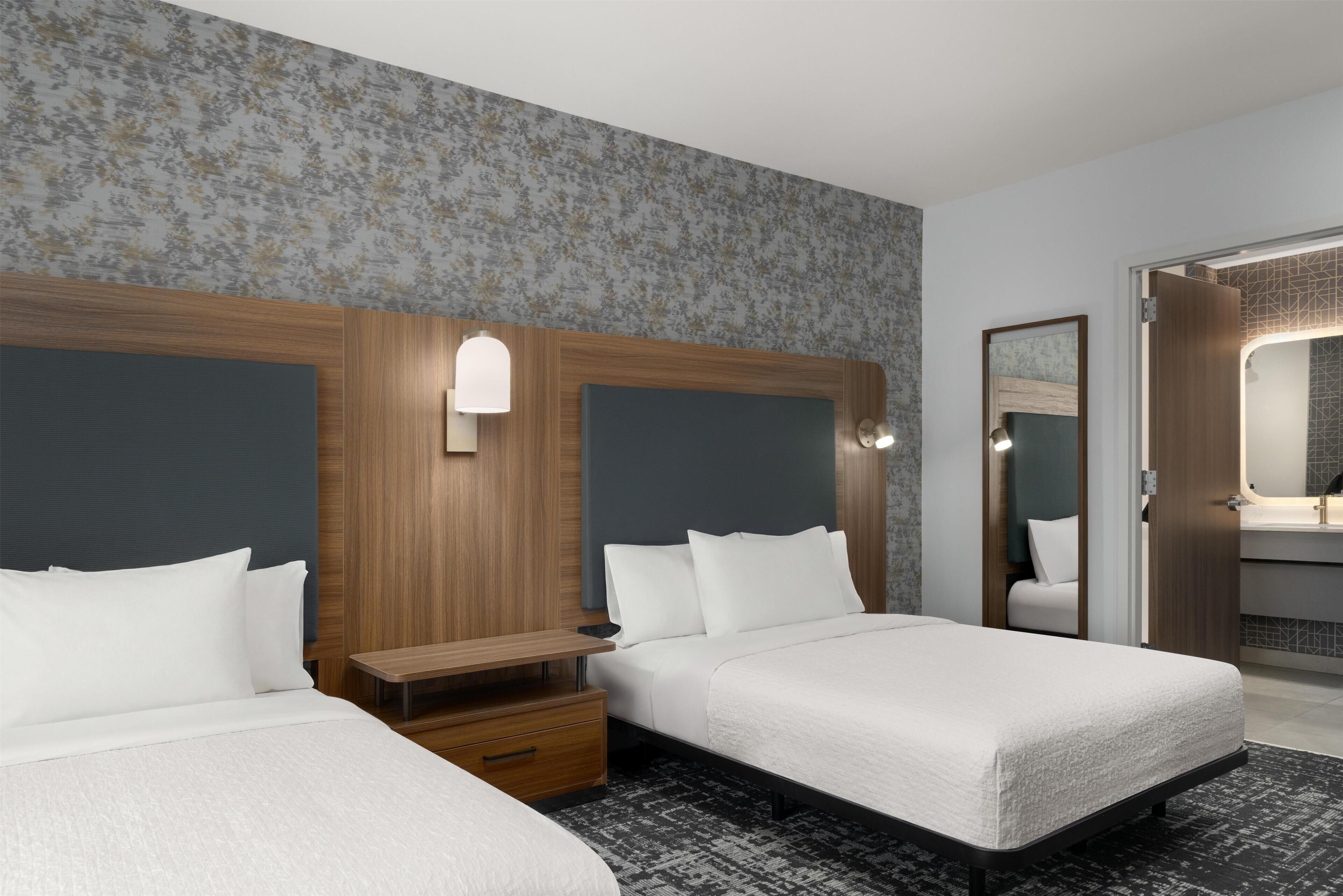 A hotel room with two beds, a wooden headboard, a nightstand, a bedside table, and a bathroom visible through an open door.