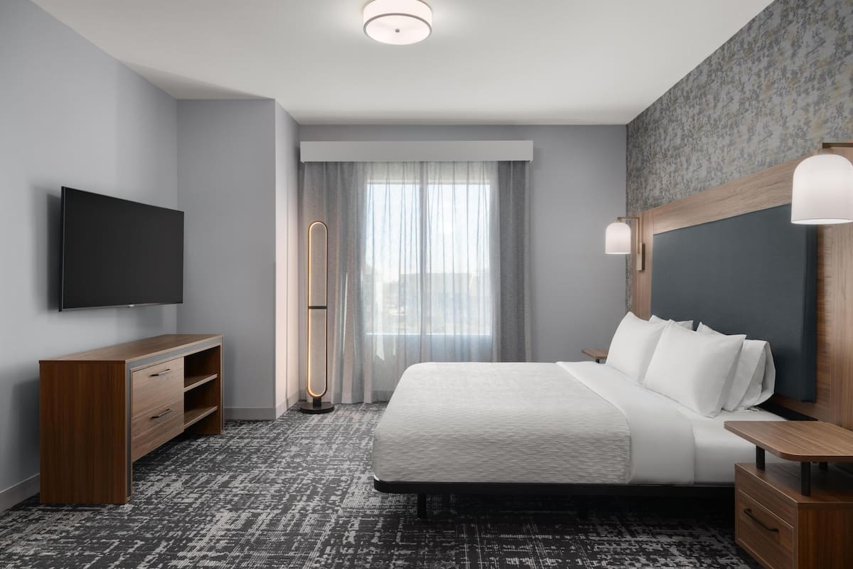 A modern hotel room with a large bed, a flat-screen TV, a wooden dresser, and a large window with sheer curtains.