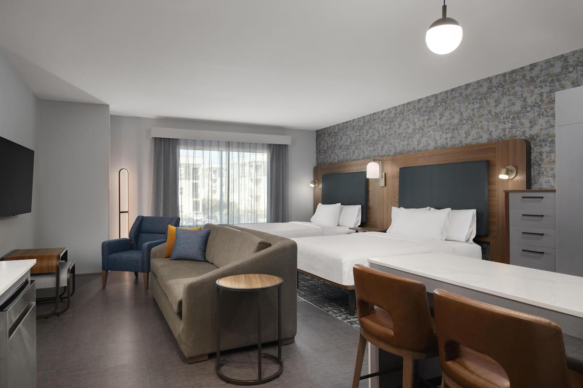 A hotel room with a sofa, two beds, a desk, a chair, a TV, and a window with curtains.
