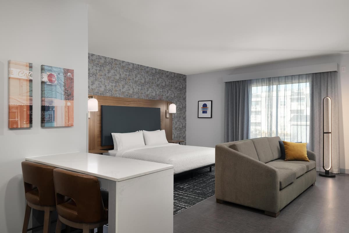 A hotel room with a bed, a sofa, a desk with chairs, a standing lamp, and artwork on the walls.