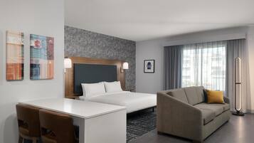 A hotel room with a bed, a sofa, a desk with chairs, a standing lamp, and artwork on the walls.