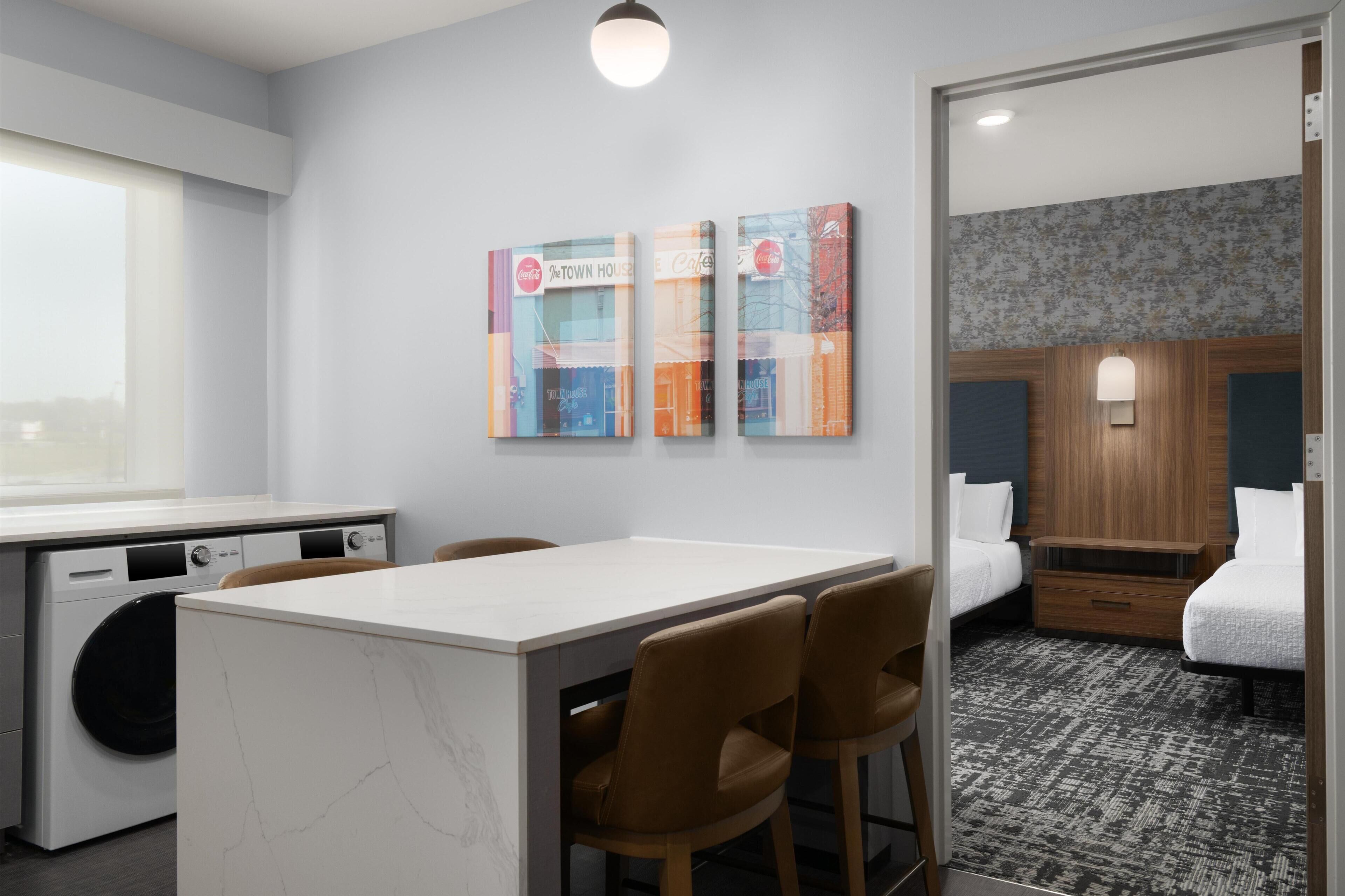 Une chambre d’hôtel avec un lit, un bureau et des chaises, un lave-linge et des œuvres d’art accrochées au mur.