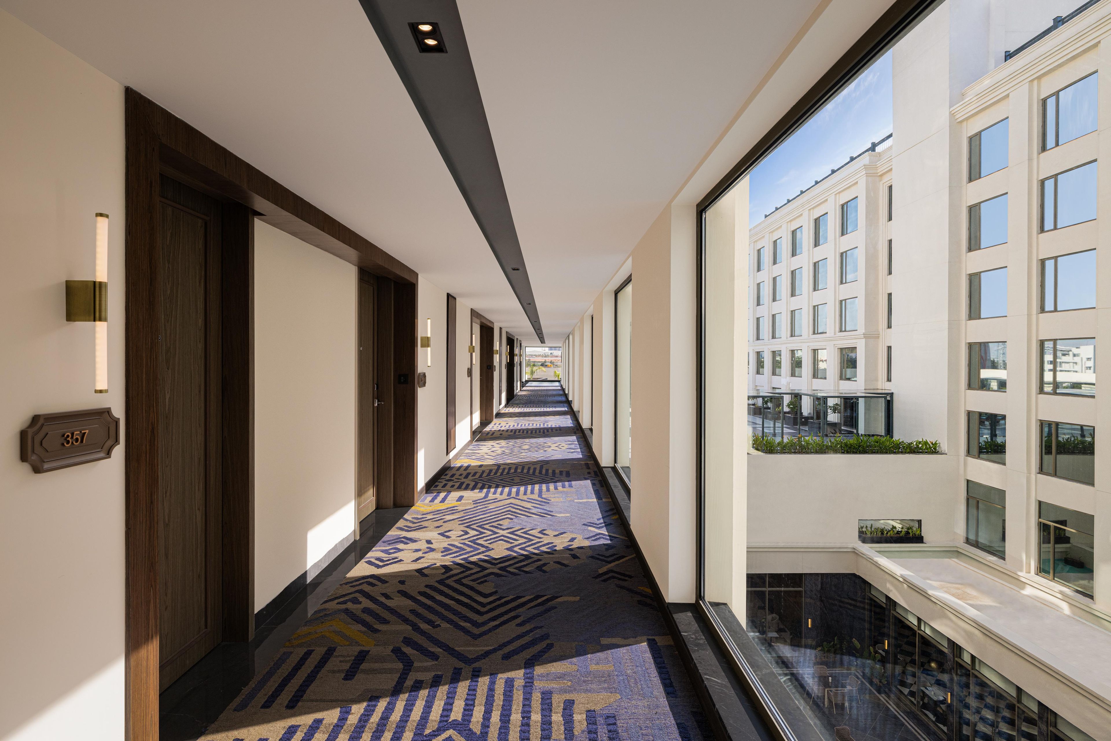 A hotel corridor with patterned carpet, a wooden door, and a view of a building with balconies.