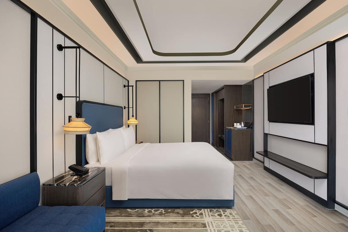 Een moderne hotelkamer met een groot bed, een aan de muur bevestigde televisie, een nachtkastje met een lamp, en een blauwe bank.