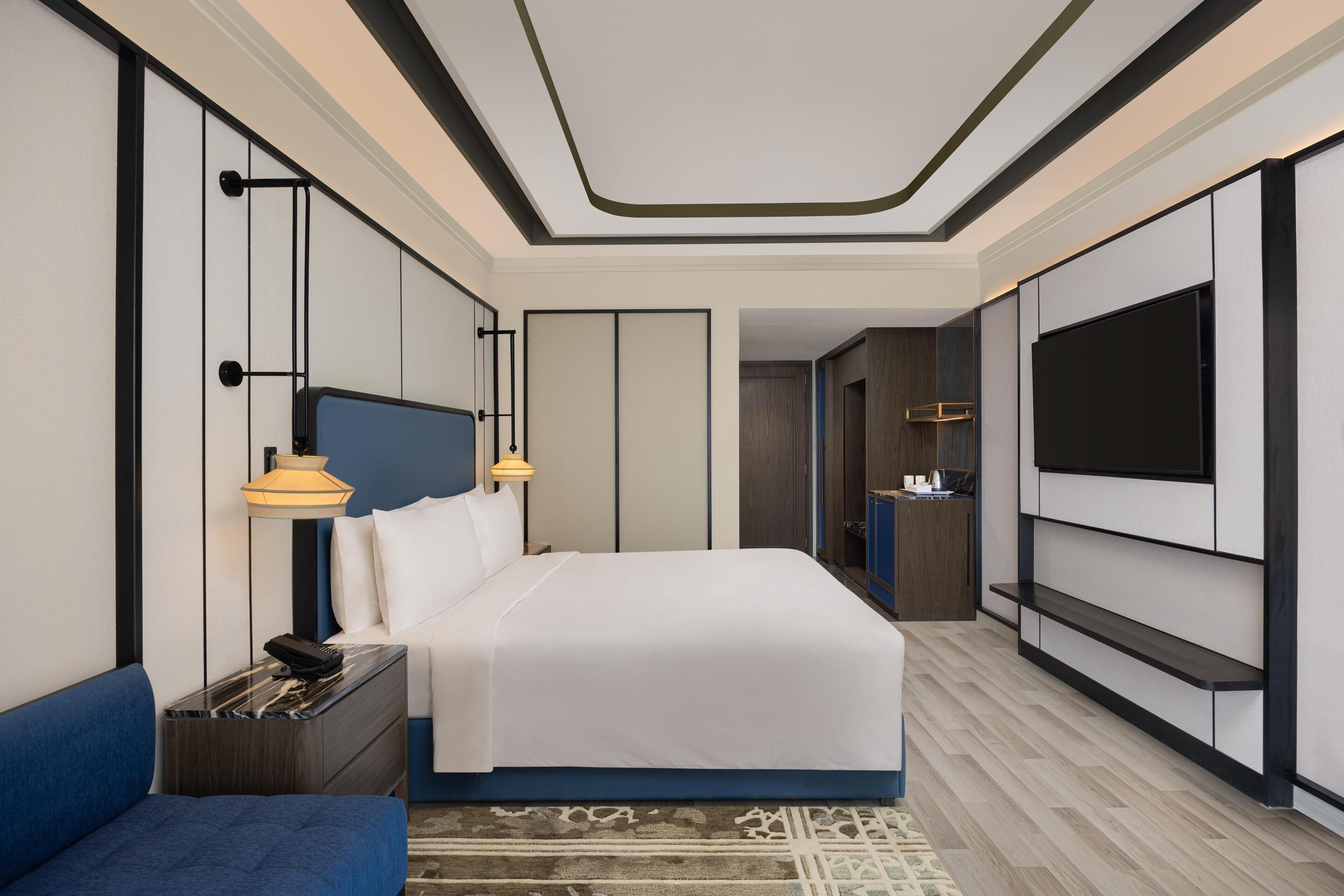 A modern hotel room with a large bed, a TV mounted on the wall, a desk with a lamp, and a blue sofa.