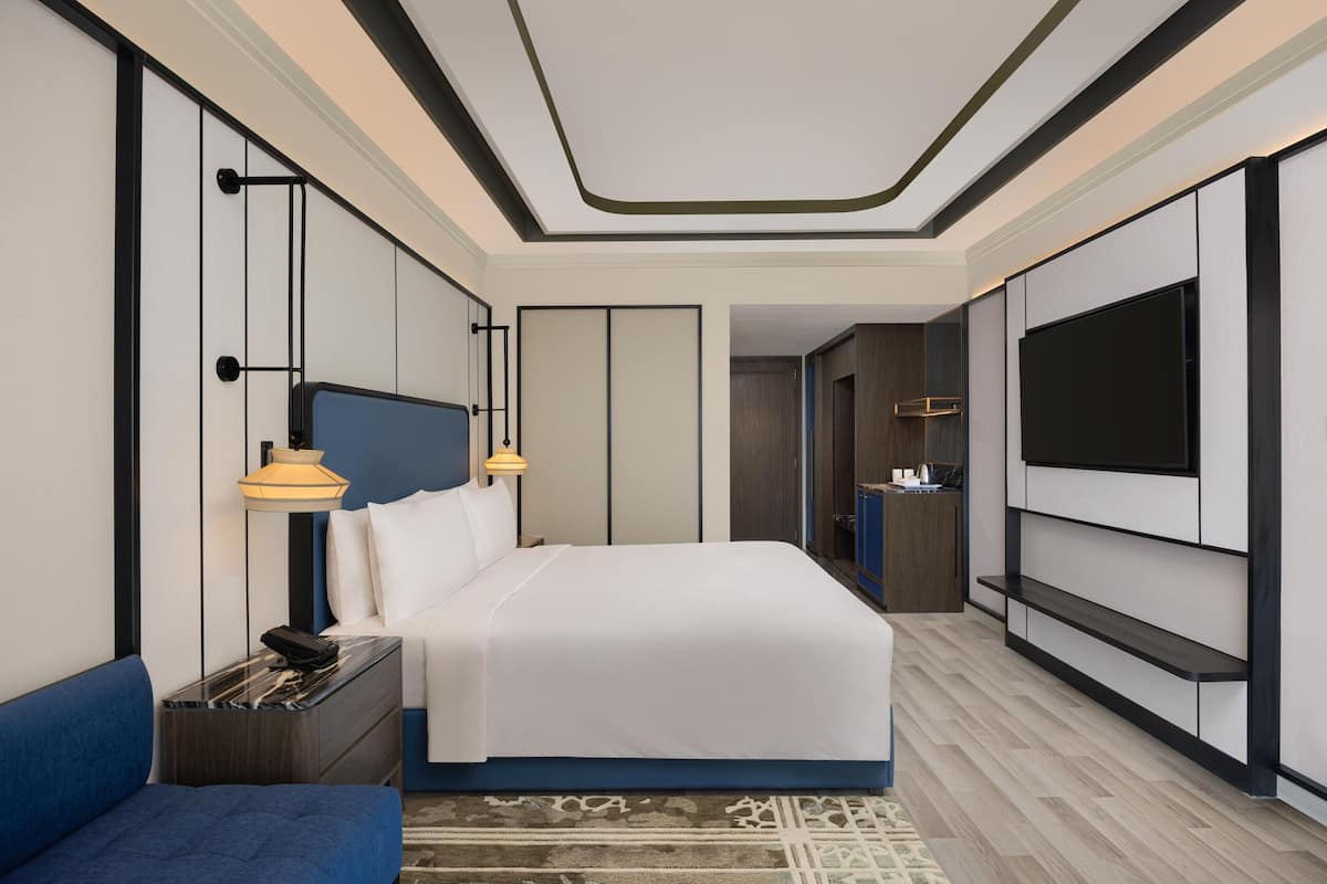 A modern hotel room with a large bed, a TV mounted on the wall, a desk with a lamp, and a blue sofa.