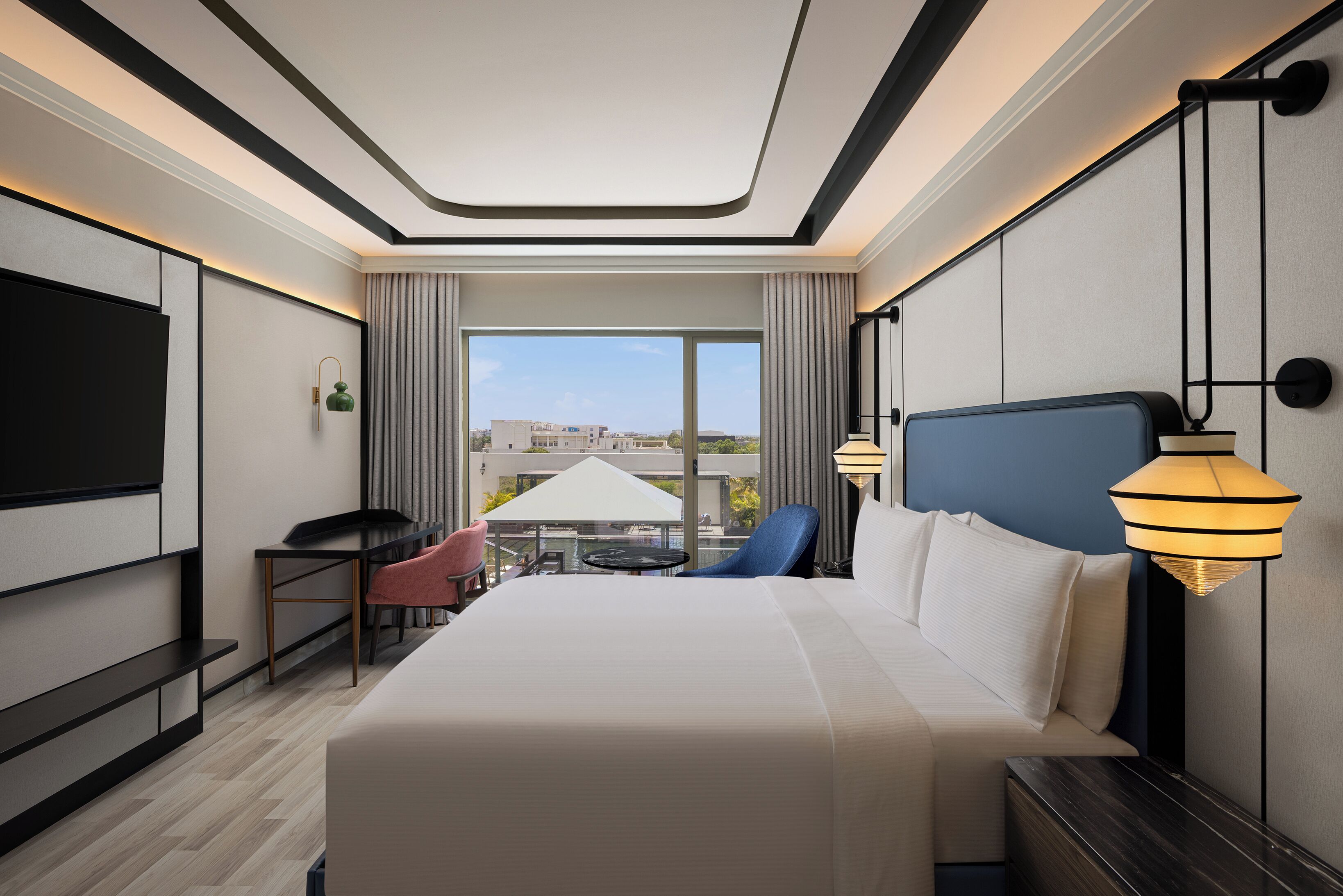 A hotel room with a large bed, a desk, a chair, a TV, and a view of the city.