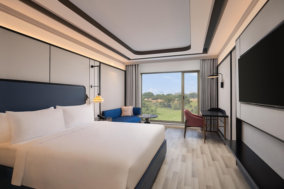 A hotel room with a large bed, a sofa, a small table, a TV, and a large window with a view.