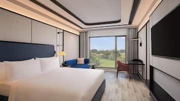 A modern hotel room with a large bed, a seating area with a sofa and table, a TV, and a large window offering a view of the outdoors.