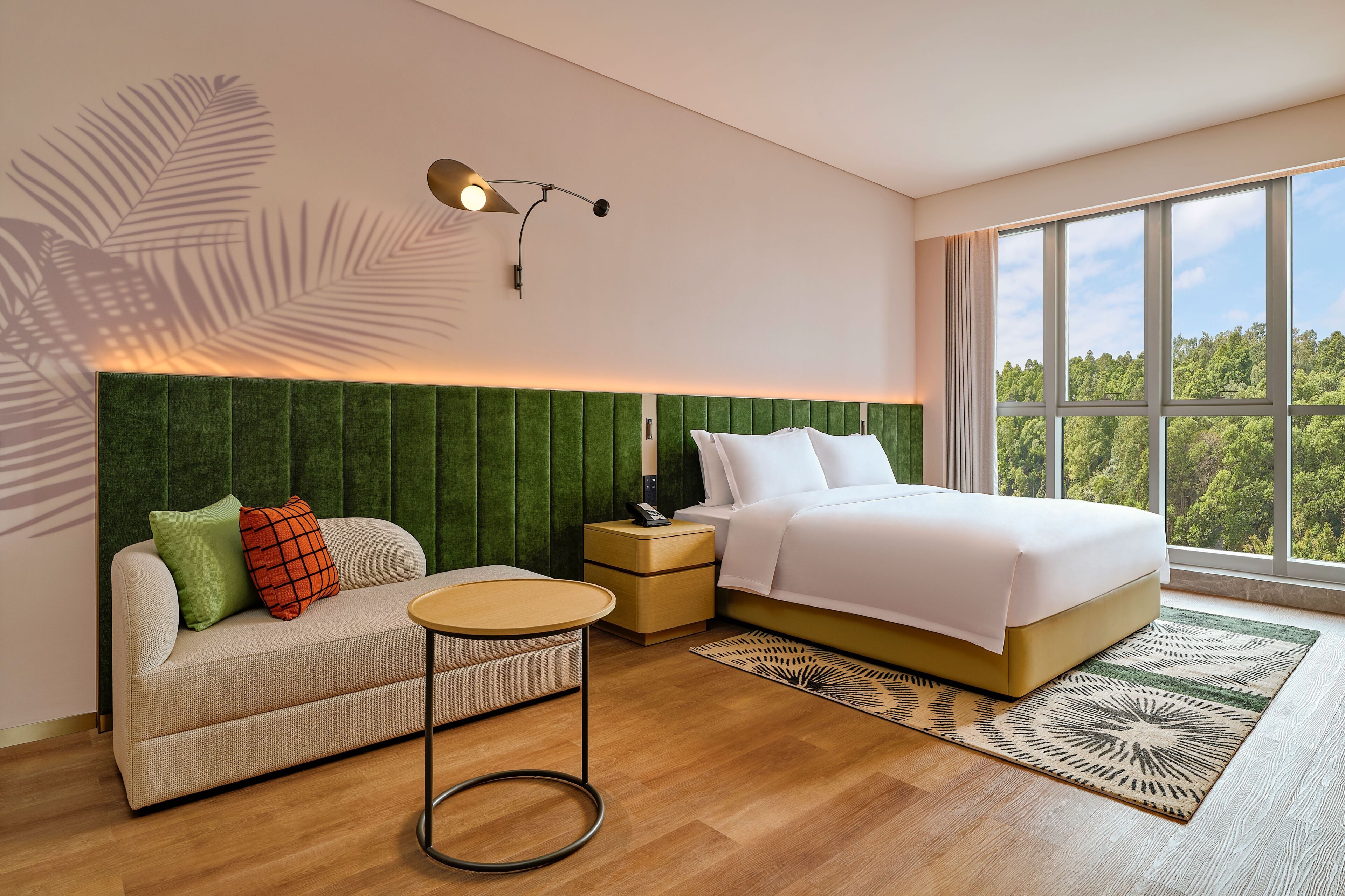 A bedroom with a bed, a sofa, a nightstand, a lamp, and a window with a view of trees.