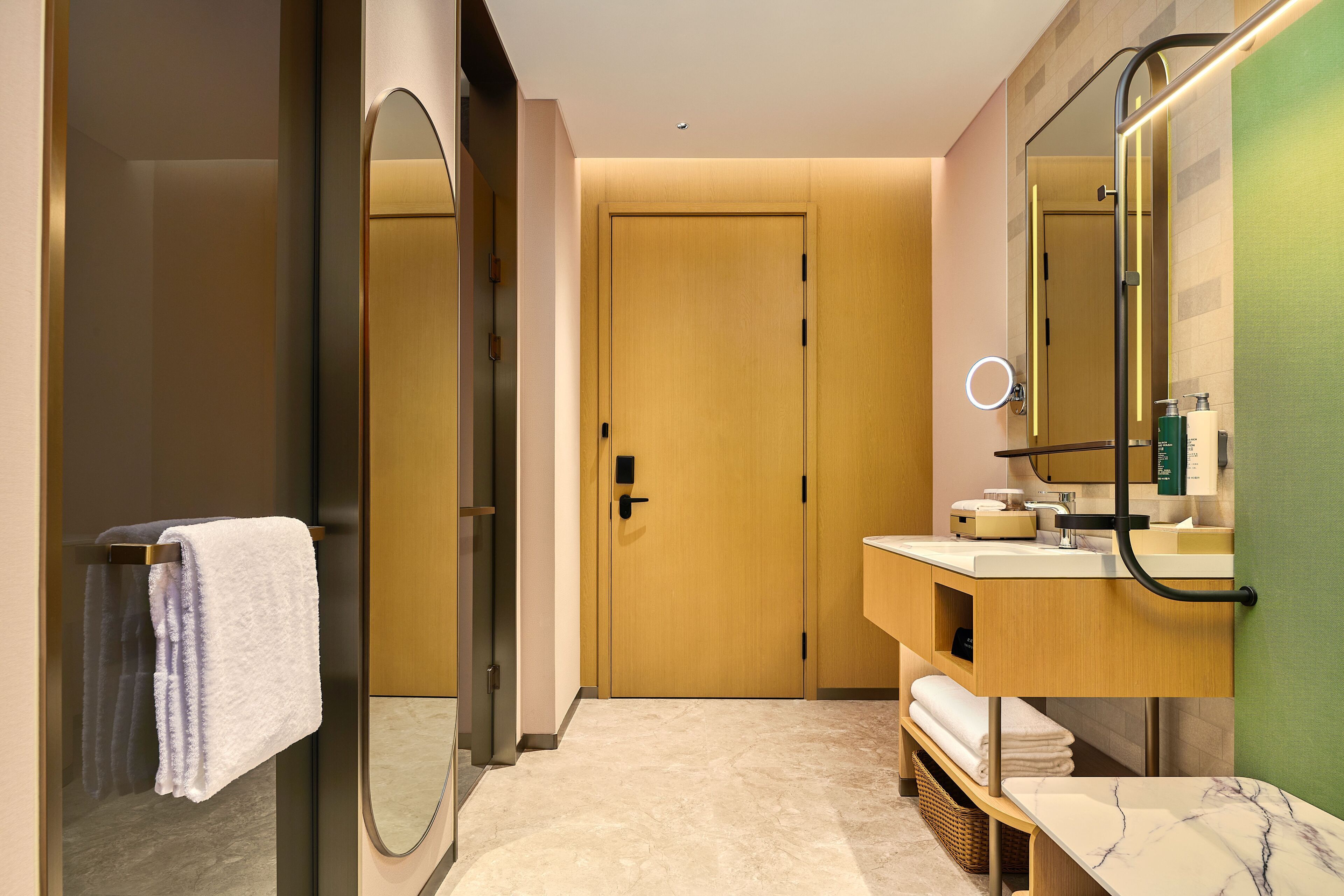 A modern bathroom with a wooden door, a glass shower enclosure, a marble countertop, and a mirror with a ring light.