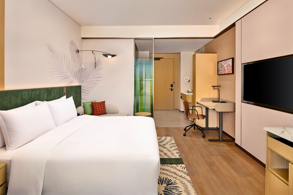 A hotel room with a bed, a desk, a TV, and a bathroom visible through a glass door.